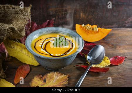Vegetable cream soup from red kuri squash with parsley garnish in a blue bowl on dark rustic wood with autumn leaves, copy space, selected focus, narr Stock Photo