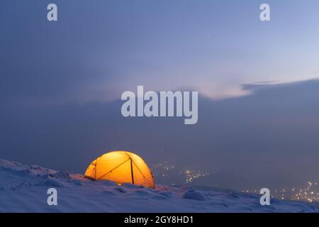 Yellow tent lighted from the inside against the backdrop of glowing city lights in fog. Amazing snowy landscape. Tourists camp in winter mountains. Travel concept Stock Photo