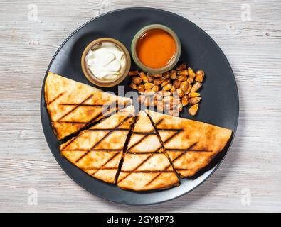 top view of cut Quesadilla from folded tortilla filled with cheese, meats, spices with sauces and fried corn seeds on plate on gray wooden board Stock Photo
