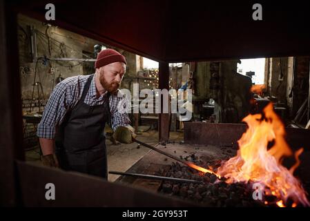 Worker in the apron burning the flame with coals in the furnace while working in blacksmith shop Stock Photo