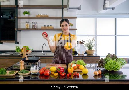 Young housewife stand smiling,  hold red and yellow bell pepper with both hands. Offering the yellow one on in the front. The kitchen counter full of Stock Photo