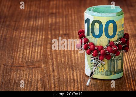 Catholic  rosary with beads and roll of Euro currency on a dark brown wooden table. Relationship of religion to money, faith, spirituality and religio Stock Photo