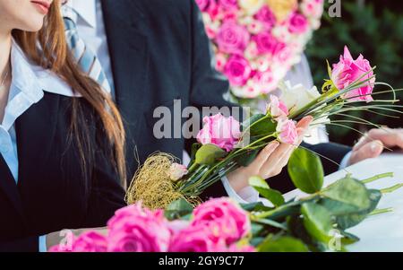 Mourning man and woman on funeral with pink rose standing at casket or coffin, a flower arrangement is in the foreground Stock Photo