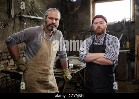 Portrait of two manual workers in aprons looking at camera while working together in the workshop Stock Photo