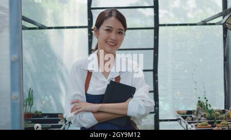 Asian beautiful young gardener woman confident with apron standing posing crossed arms and smiling to camera at greenhouse cactus plant nursery, small Stock Photo