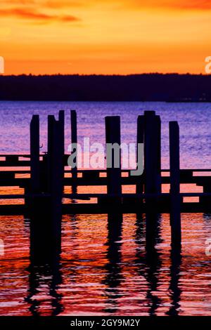 Dock silhouettes in a river of purple red and orange with a yellow and orange sky and a tree line against the sky Stock Photo