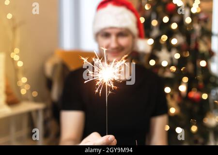 Young man in Santa hat is holding burning sparkler near Christmas tree. Christmas lights in guy hands on blurred background with decorated Christmas t Stock Photo