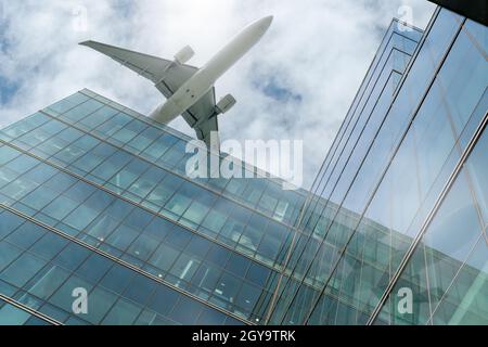 Airplane flying above modern office building. Exterior facade of skyscraper building. Business trip. Reflection in transparent glass windows. Aviation Stock Photo