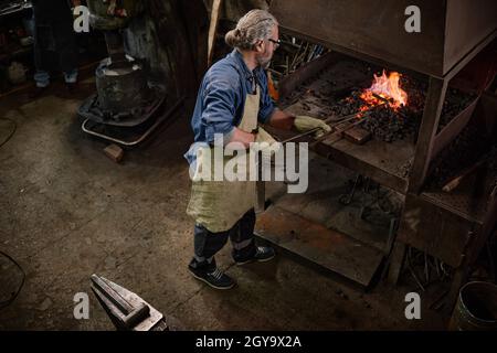 Worker putting coal into furnace for making details from iron in the workshop Stock Photo