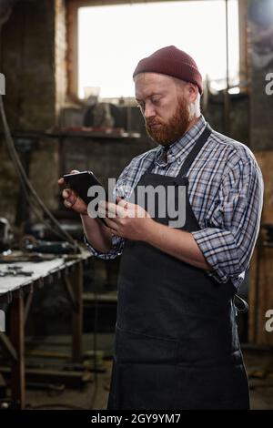 Mature engineer in apron typing on digital tablet during his work in workshop Stock Photo