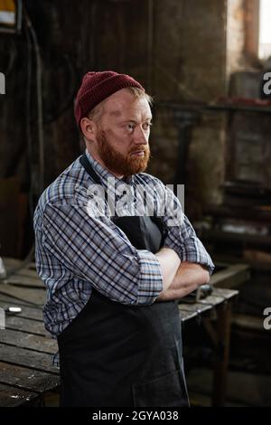 Mature man in apron standing with arms crossed and thinking about something in the workshop Stock Photo