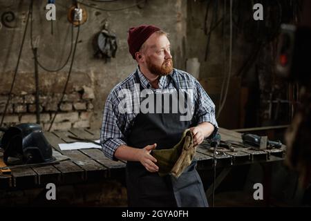 Mature blacksmith in apron holding protective gloves standing near his workplace in the workshop Stock Photo