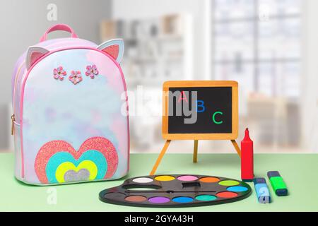 Back to school. Colorful school equipment, a bright school backpack and a blackboard with ABC letters on green table over an abstract blurred classroo Stock Photo