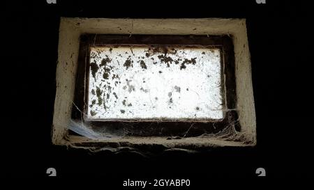 a small rectangular dirty and dusty web window in a large basement. White light penetrates the dark basement Stock Photo