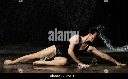 young woman with black hair lies on the ground under raindrops on a black background. Woman dressed in black bodysuit Stock Photo