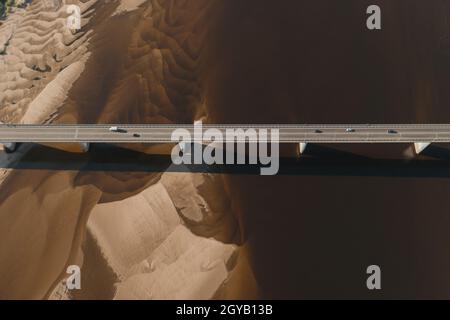 Aerial drone shot of cars driving on bridge over river and sandy riverside at sunset. Amazing aesthetic top view shot with beautiful coast with dune s Stock Photo