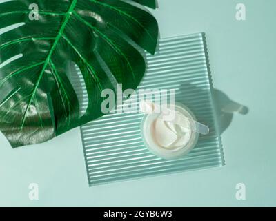 Cosmetic cream or moustirizer on transparent ribbed acrylic plate, tropical palm monstera leaf over blue background. Open round jar, aesthetic swirls Stock Photo