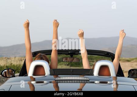 Back view portrait of two women raising arms in a convertible car enjoying views in the mountain Stock Photo