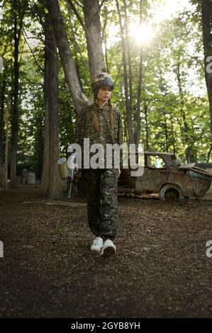 Female paintball player with gun poses on playground in the forest. Extreme sport with pneumatic weapon and paint bullets or markers Stock Photo