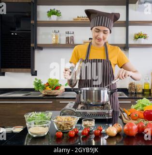 Young asian housewife dressed in an apron and a hair cap, preparing  the meat stew in a modern kitchen. Stock Photo