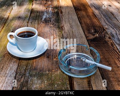 cigarette cup A cup of coffee and a glass ashtray with a cigarette on a wooden table.  Coffee drink. A cigarette in an ashtray. Wooden table. Hot drink. Street  Cafe Stock Photo