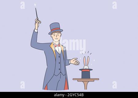 Magician making trick during work concept. Young smiling man magician wearing traditional costume standing with stick making magic trick with rabbit i Stock Photo