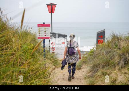 Rear view of a woman walking between sand dunes to a beach in Wales with the sea and warning signs ahead of her.