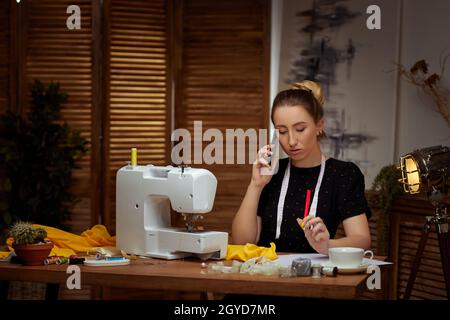 Beautiful smiling young seamstress working on sewing machine and talking by phone Stock Photo