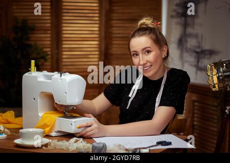 Beautiful smiling young seamstress sews clothes on sewing machine at home Stock Photo