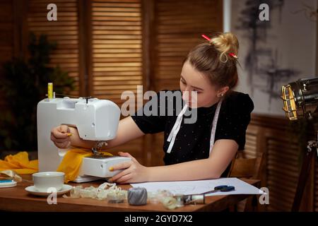 https://l450v.alamy.com/450v/2gyd7x2/beautiful-young-seamstress-with-measuring-tape-on-neck-sitting-at-working-table-and-working-on-sewing-machine-2gyd7x2.jpg