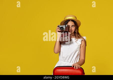 stylish girl with red suitcase holding retro vintage photo camera, doing photo shot on yellow background. teenager is ready for summer travel Stock Photo