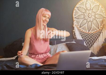 Smiling woman in elegant pink pajamas checks time on smartwatch in bedroom Stock Photo