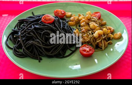 black spaghetti on a green plate accompanied by prawns, gluttony and tomato on a pink tray Stock Photo