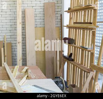 A desk full of wood piles. Wood planks of various sizes lean on a white brick wall. Atmosphere in the workshop room. Stock Photo