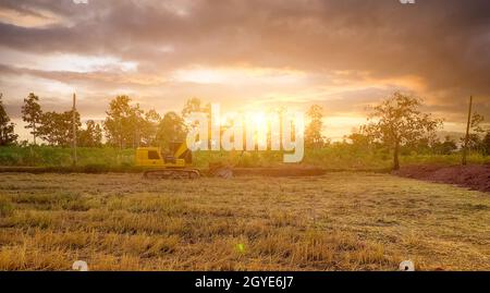 Landscape of rice field and tropical forest with sunset sky. Backhoe working by digging soil. Excavator digging on soil at rice field after harvest in Stock Photo