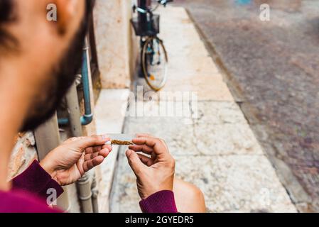 A young man uses cigarette paper to roll a cigarette on a street. Stock Photo
