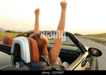 Two women in a convertible car driving and raising arms in a mountain road Stock Photo