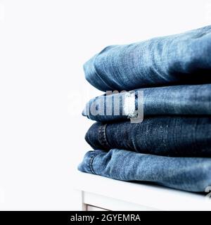 Stack of different blue denim jeans trousers on the shelf or table. Photo of stacked various shade jeans close up on white background with copy space Stock Photo