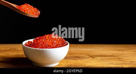 Red caviar in a wooden cup on a black background with a spoon. A large pile of bright caviar. Fresh delicious caviar. A place for advertising, logo, l Stock Photo