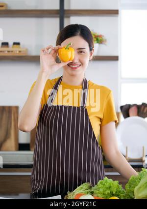 Young housewife dressed in an apron stand smiling, hold yellow bell pepper in eye position. Stock Photo