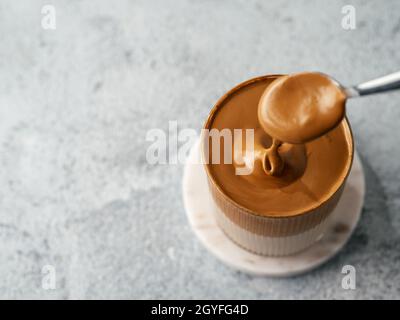 Trendy korean drink - dalgona coffee. Glass jar with cold or hot beverage - whipped instant coffee with milk poured from spoon. Gray cement background Stock Photo