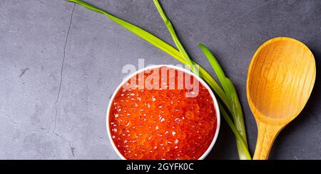 Red caviar in a wooden cup on a gray background with a spoon and green onions. A large pile of bright caviar. Fresh delicious caviar. A place for adve Stock Photo