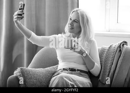 Beautiful mature woman with cup of coffee taking selfie at home