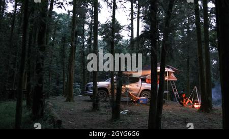 Camper enjoying the alternative vacation - car with tent on the roof and alone man cooking in the forest. Adventure travel lifestyle wanderlust Stock Photo