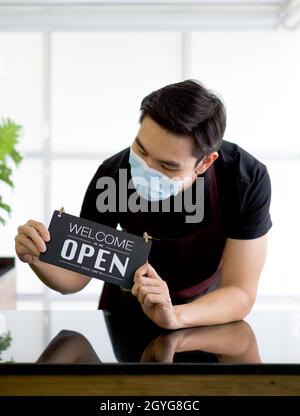Young asian man with a face mask holds an opening sign in front of a coffee shop counter. Stock Photo