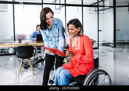 Mexican transgender woman in a wheelchair with young office colleague sharing information at the workplace in Latin America Stock Photo