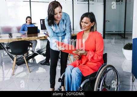 Mexican transgender woman in a wheelchair with young office colleague sharing information at the workplace in Latin America Stock Photo