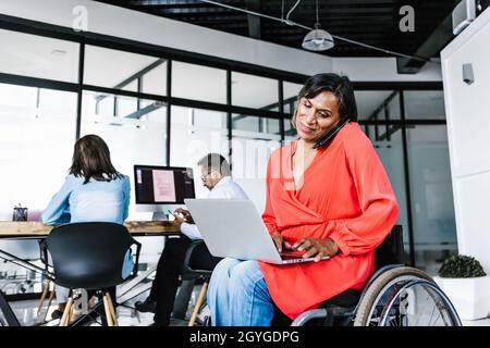 Mexican transgender businesswoman in wheelchair making phone call while working on computer at work in Mexico Stock Photo