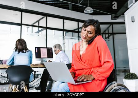 Mexican transgender businesswoman in wheelchair making phone call while working on computer at work in Mexico Stock Photo