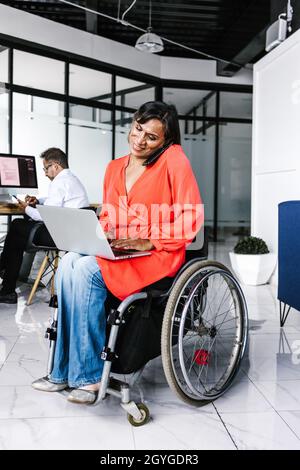 Latin transgender woman sitting in wheelchair using computer while talking on the phone in office in Mexico Stock Photo